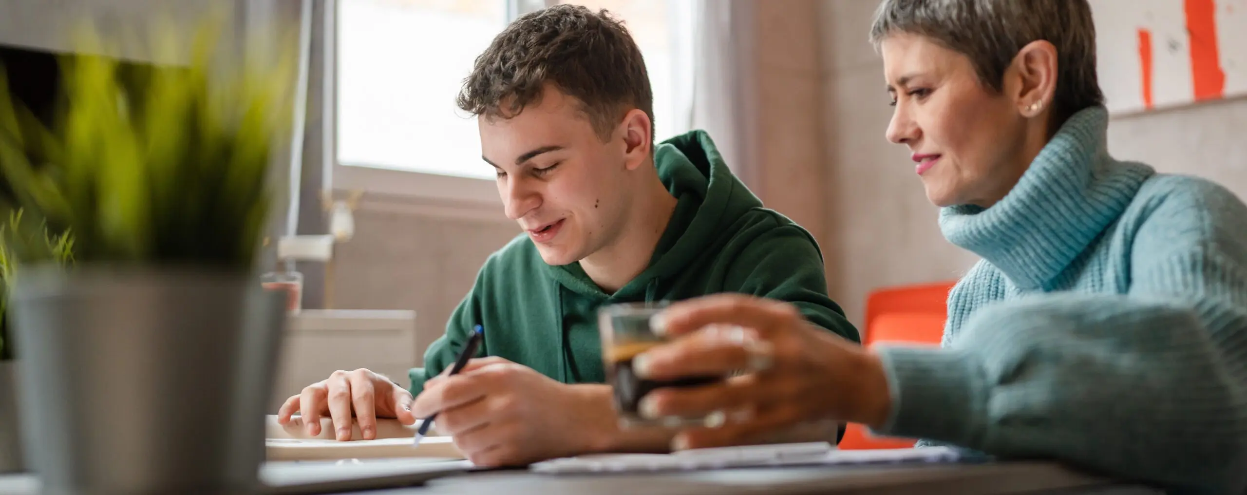 A mother drinking coffee is sitting next to he son at a computer in their home. She is helping with his homework as they both look down at it.