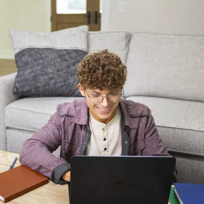 A student smiling and working on his laptop in his home.