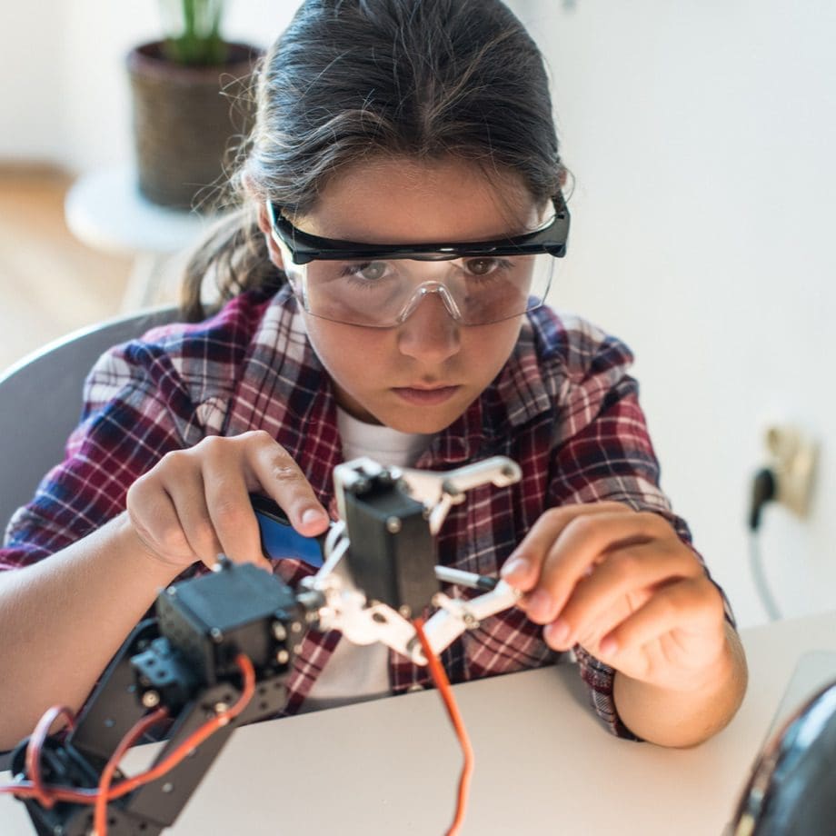 Young student in safety glasses is concentrating while working with tools on a small part of a robot.