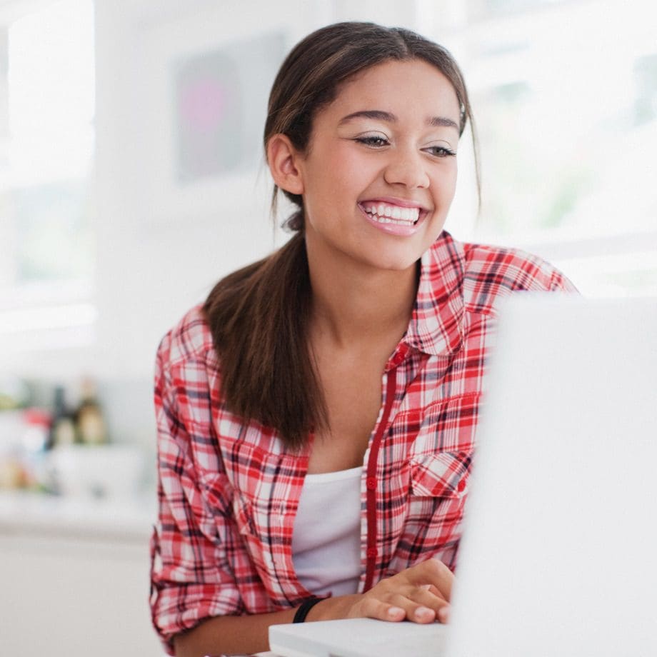 High school student in bright room is working on laptop with a huge smile on her face.