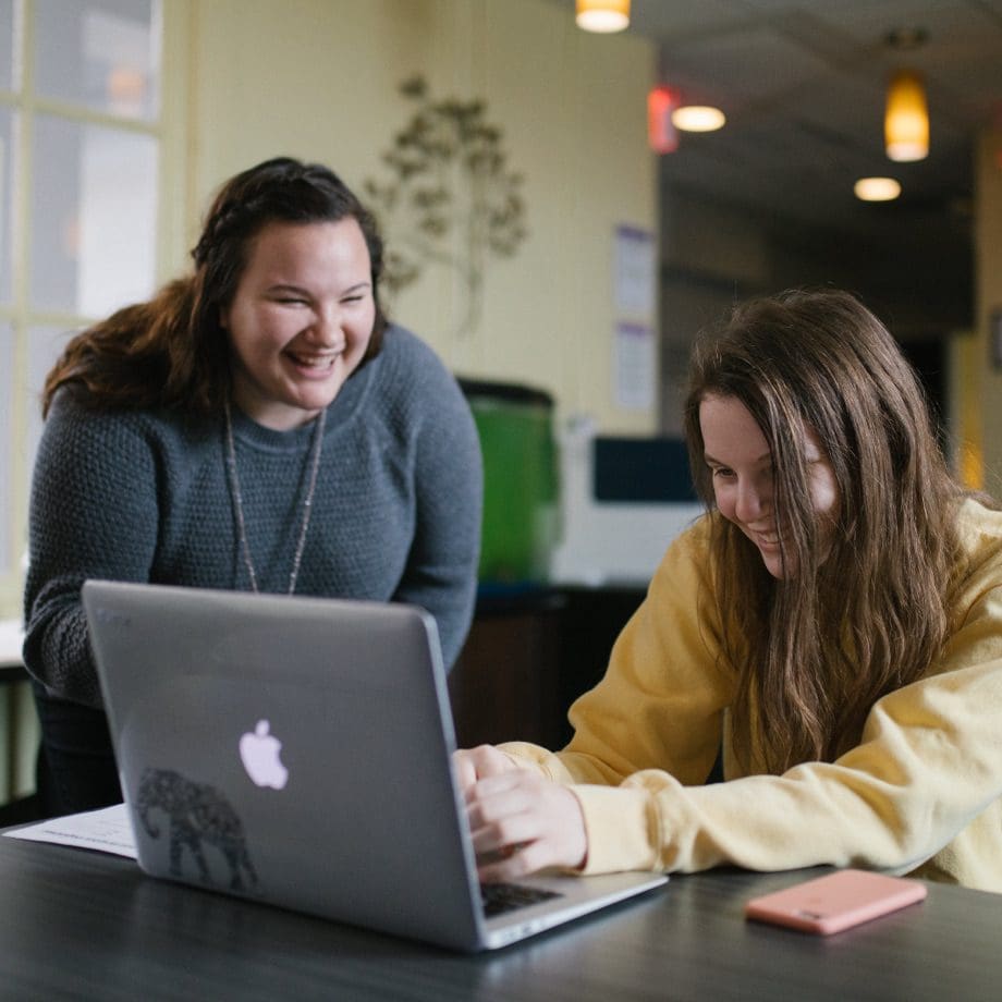 A student is smiling and typing on her laptop, while a happy teacher is looking over her shoulder and helping.