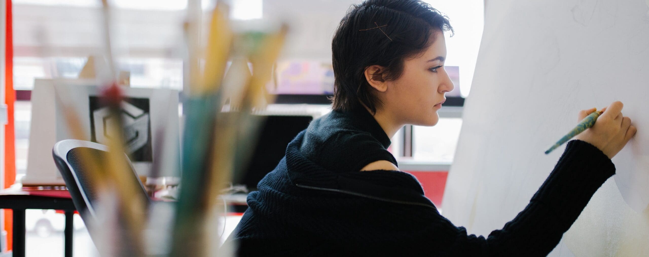 An art student is focused on painting at a large easel. They are in an art room with paintbrushes in the foreground.